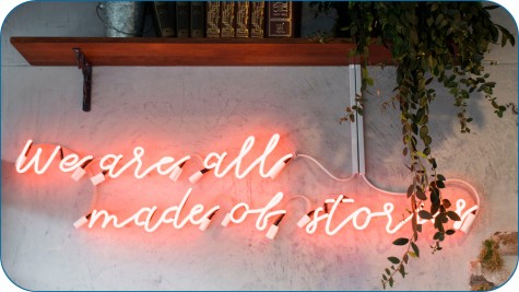 Dieses Bild ist ein Teaserbild und für das Verständnis des Artikels irrelevant.
Die Abbildung zeigt eine dekorative Neon-Leuchtschrift an einer Wand.
Die Schrift leuchtet in einem hellen Rosa-Orange und lautet: „We are all made of stories“.
Darüber befindet sich ein Holzregal mit alten Büchern und einem Metallgefäß.
Rechts hängt eine grüne Pflanze mit langen herabhängenden Ranken, die teilweise die Leuchtschrift überdecken.
Die Wand im Hintergrund wirkt grau und leicht strukturiert.