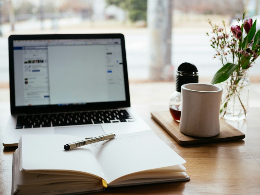 Schreibtisch-Szene mit einem geöffneten Laptop, einem Notizbuch samt Stift, einer Tasse Kaffee und einem Blumenstrauß in einem Glasgefäß – sinnbildlich für konzentriertes Arbeiten oder Lernen in einer angenehmen Atmosphäre.